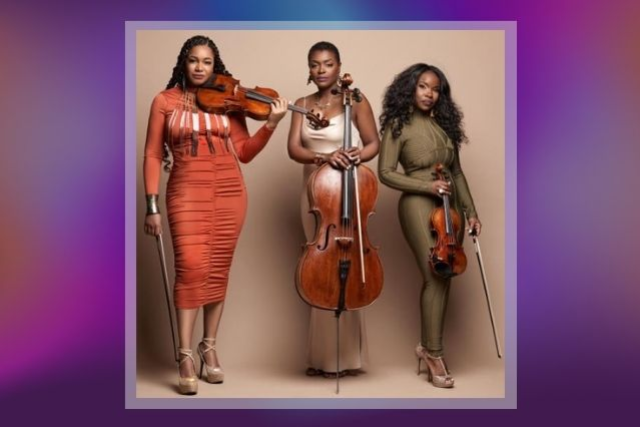 Three women stand holding musical instruments, from left to right, holding a viola, a cello, and a violin.